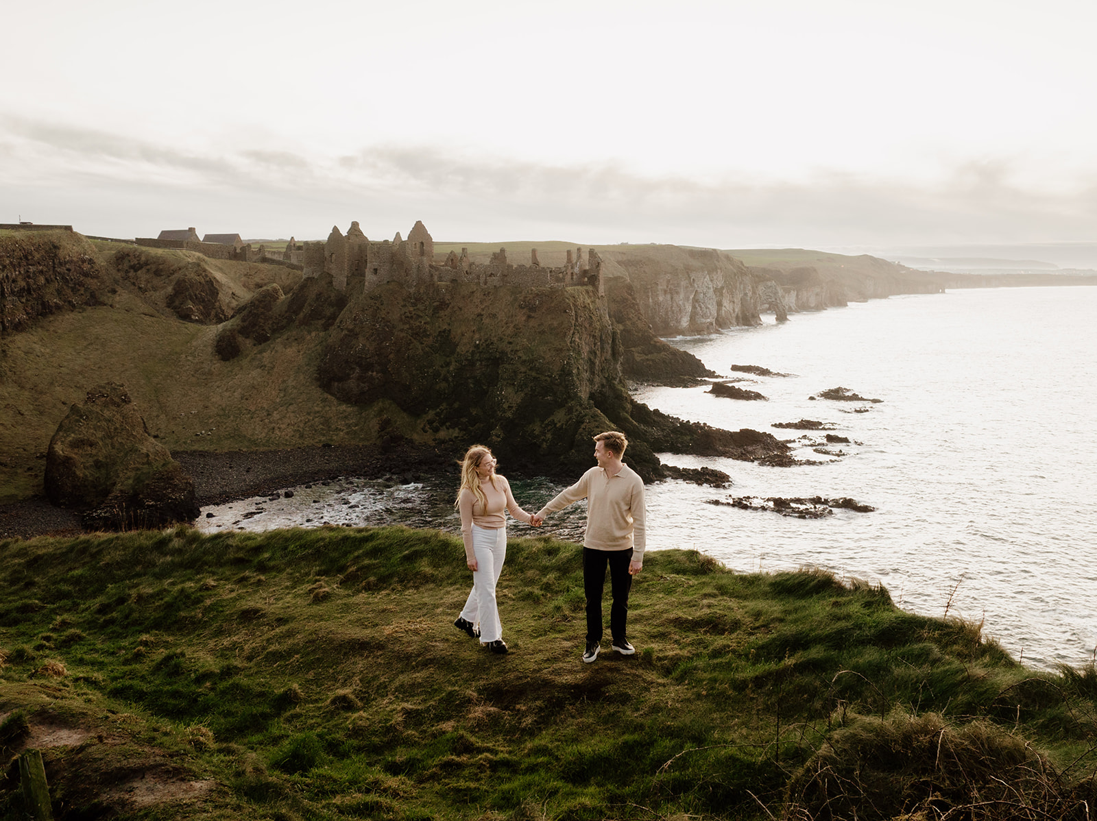 couple walking at the edge of the cliff with dunlice castle in the background for their pre engagement shoot