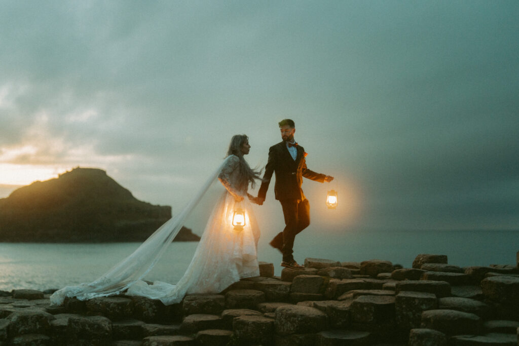 A bride and groom holding lanterns as they walk across the stones at Giants Causeway. Bride has a long veil