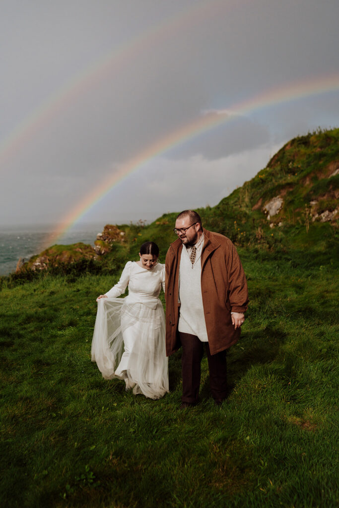 A couple walking in the rain on green grass at Kinbane Castle Ireland with a double rainbow behind them.